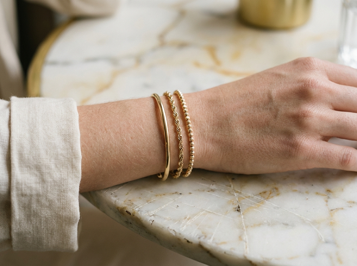 Bracelets fins dorés empilés sur un poignet de femme posé sur du marbre blanc, vue rapprochée